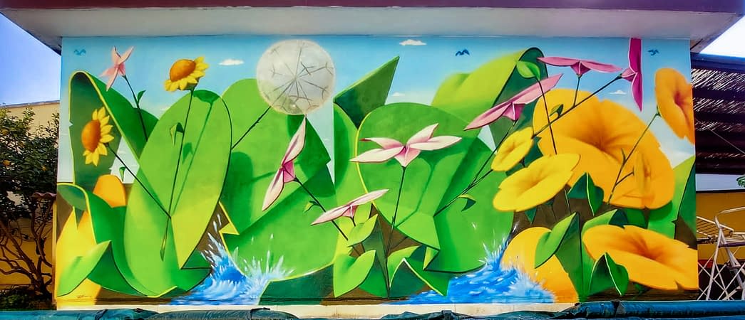 Total view of a large mural painting depicting big green leaves with geometric rounded shapes, composing the tag Dado, and big yellow-orange flowers along with smaller pink and light yellow flowers floating on deep blue water. Artwork by Dado