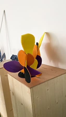 Side angle view of a sculpture made of thin bent layers with a soft felt finish in a mix of high saturated yellow, orange, purple and black colors. Artwork by Dado