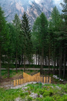 Panoramic view of a mountain landscape with a series of tall wooden boards planted in the ground forming a big X in the middle of a mountain path, in front of a grass field, with high trees in the background. Sculpture installation by Dado.