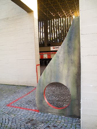 Sculpture representing an almost flat "D" sinuous letter installed next to a concrete wall. The red outline of the letter extends from the artwork and runs on the ground. Close view