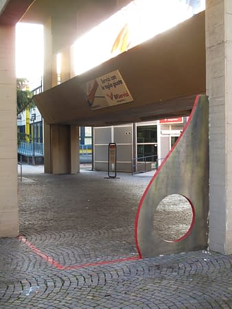 Sculpture representing an almost flat "D" sinuous letter installed next to a concrete wall. The red outline of the letter extends from the artwork and runs on the ground. Front left view