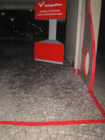 Sculpture representing an almost flat "D" sinuous letter installed next to a concrete wall. The red outline of the letter extends from the artwork and runs on the ground