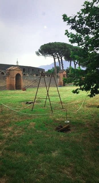 A modern sculpture installation consisting of an old iron swing with the seat still suspended in the air, in the middle of a grass field with an ancient long brick wall in the background . Sculpture by Dado