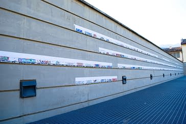 A long gray concrete wall with three long lines of paper sheets with drawings, each one depicting a train wagon with a graffiti on the side, are stitched together to form al long train