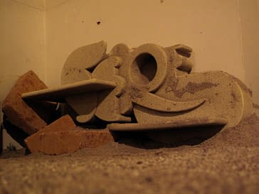 A grey, rock-style, sculpture formed of letters in different styles which composing the name "Sere". The sculpture is laying on the floor along with some thrown brick. Artwork by Dado