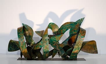 Front view of a sculpture made of copper ribbons sinuously coiled, forming the tag of the artist Dado. The copper colored surface is texturized with extensive green oxidation stains.