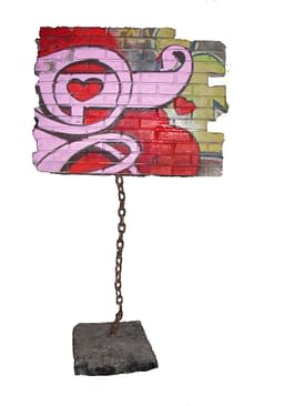 A modern art sculpture consisting in a square piece of a brick wall painted with graffiti floating off the ground trying to fly away but a strong heavy chain keep it connected to the ground. Artwork by Dado