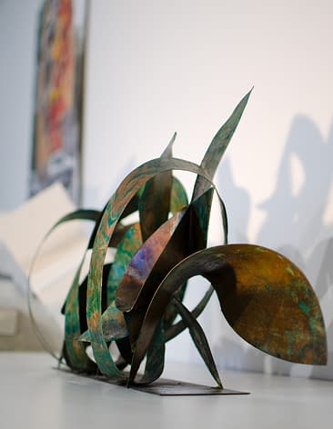 Side view of a sculpture made of copper ribbons sinuously coiled, forming the tag of the artist Dado. The copper colored surface is texturized with extensive green oxidation stains.