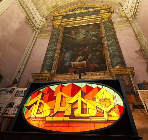 Inside an old church is placed for display a stained glass window made colored glass pieces representing the word Dado in a graffiti style at the bottom of stylized skyscrapers. bottom right view