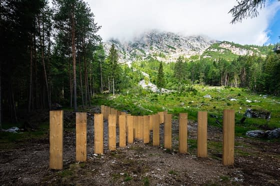 Closer view of tall wooden boards planted in the ground, in the middle of a mountain path, forming a big X. High mountains landscape in the background. Sculpture installation by Dado.
