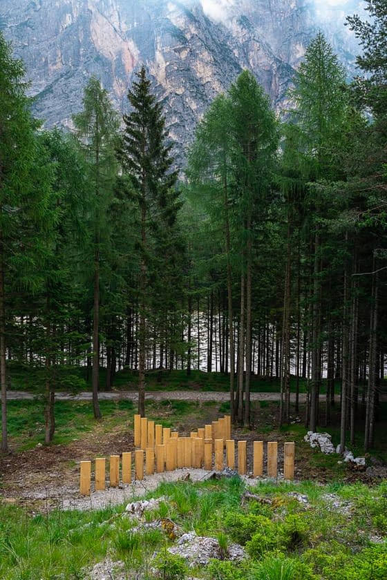 Panoramic view of a mountain landscape with a series of tall wooden boards planted in the ground forming a big X in the middle of a mountain path, in front of a grass field, with high trees in the background. Sculpture installation by Dado.