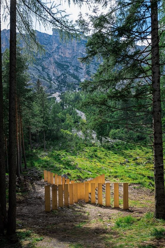 Panoramic view of a mountain landscape with a series of tall wooden boards planted in the ground forming a big X in the middle of a mountain path with high mountains in the background. Sculpture installation by Dado.