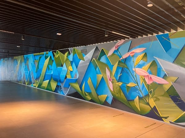 Long wall decorated with intersecting blue and green ribbons and a bunch of delicate pink flowers appearing on the right. Aerosol art by Dado.