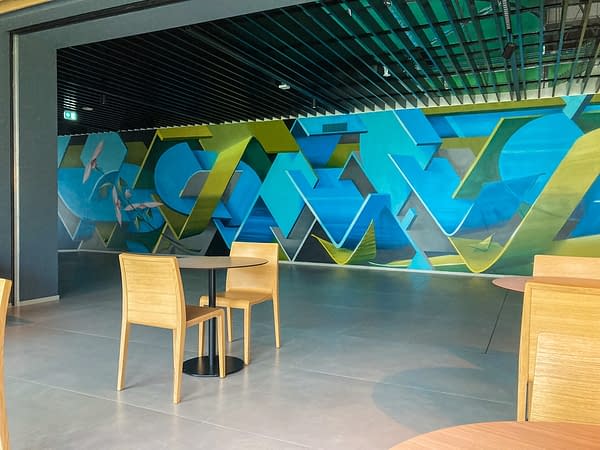 Long wall decorated with intersecting blue and green ribbons in the back of an open space hall of the EVO Bonfiglioli Branch in Bologna. Aerosol art by Dado.