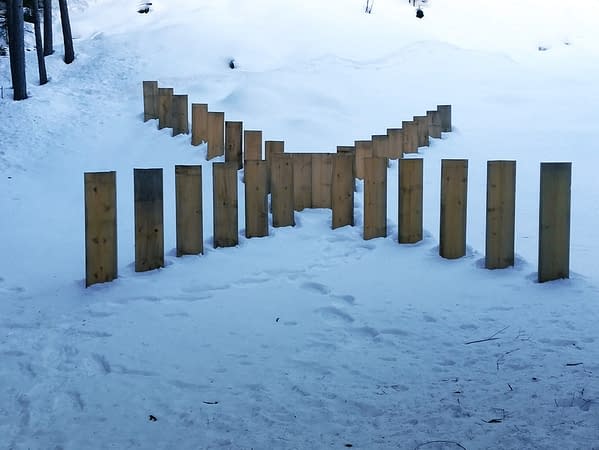 Front view of a wood sculpture composed from a series of tall wooden boards planted in the ground forming a big X just in the middle of a mountain path. The ground covered by the snow.