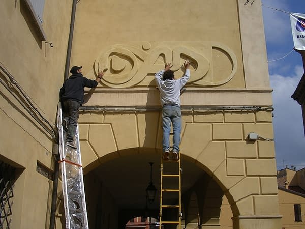 The artist Dado Ferri and his helper on a work ladder applying the bas-relief Sculpture named Lettering Medioevale on a wall for the Exhibition
