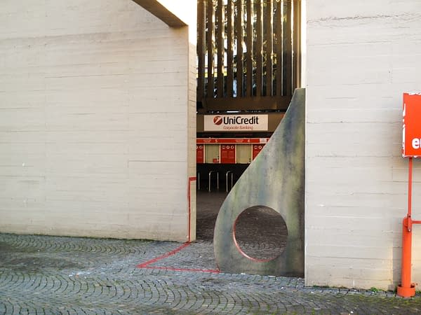 Sculpture representing an almost flat "D" sinuous letter installed next to a concrete wall. The red outline of the letter extends from the artwork and runs on the ground. Left view