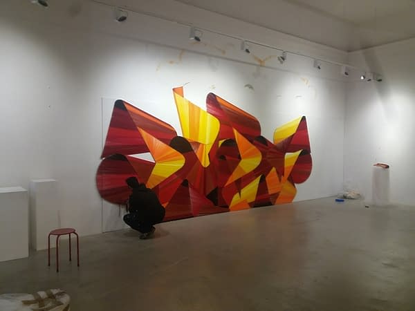 A wool made graffiti in the tones of yellow, orange and various reds lying on a white wall inside a gallery. Artworks by Dado