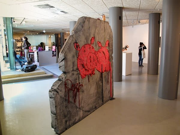 A big rock sculpture with two wool made flat pig characters stitched in the middle, inside an art gallery. Artwork by Dado