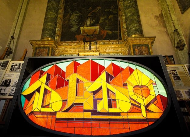 Inside an old church is placed for display a stained glass window made colored glass pieces representing the word Dado in a graffiti style at the bottom of stylized skyscrapers. Front view