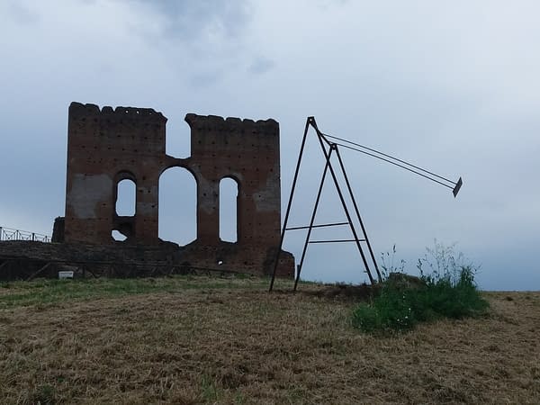 A modern sculpture installation consisting of an old iron swing with the seat still suspended in the air with an ancient roman wall ruin in the background. Sculpture by Dado