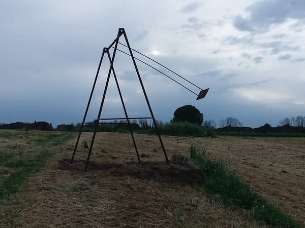 A modern sculpture installation consisting of an old iron swing with the seat still suspended in the air in the middle of a grass field. Sculpture by Dado