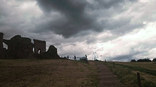 Wide view of an hill, on an overcast day, on top of which are located an old roman ruin and a modern art installation consisting of an old iron swing with the seat still suspended in the air. Installation by Dado