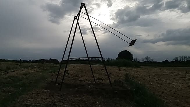Wide view of a field on top of an hill, on an overcast day, in the middle of which is placed a modern art installation consisting of an old iron swing with the seat still suspended in the air. Installation by Dado