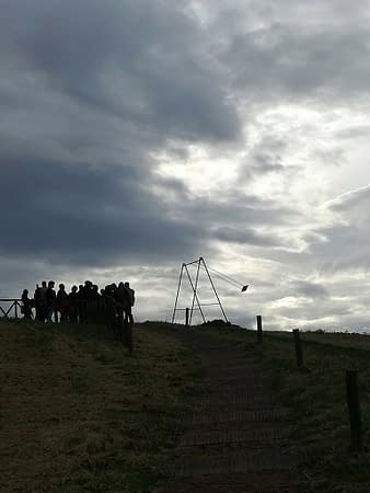 Wide view of the top of an hill, on an overcast day, on top of which a group of people is admiring a modern art installation consisting of an old iron swing with the seat still suspended in the air. Installation by Dado