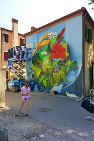 Dado painting a Graffiti-mural spherical lettering with multi colored flowers-ground view of the entire wall and the artist on an industrial elevator while a kid in pink dress is watching