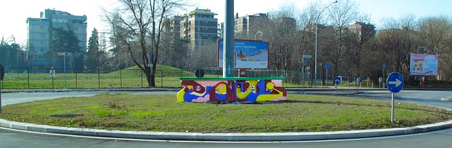 A graffiti style bright colored sculpture in the middle of a traffic circle in a public road path. Installation by Dado