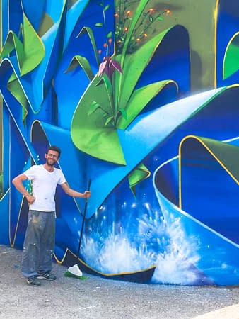 Dado Ferri posing in front of his freshly finished mural artwork depicting wide blue ribbons decorated with purple flowers and green leaves.