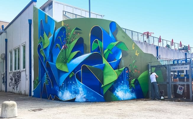 Wide location view of a large mural painting depicting wide sinuous blue ribbons decorated with purple, red and yellow flowers and green leaves and water splashes. Artwork by Dado