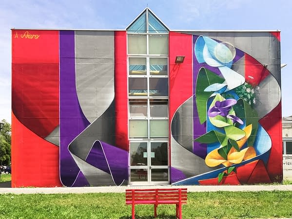 Front view of an high school building face with a mural painting depicting wide gray and purple ribbons composing the name Piero in a graffiti wildstyle decorated with yellow and purple flowers. Artwork by Dado.