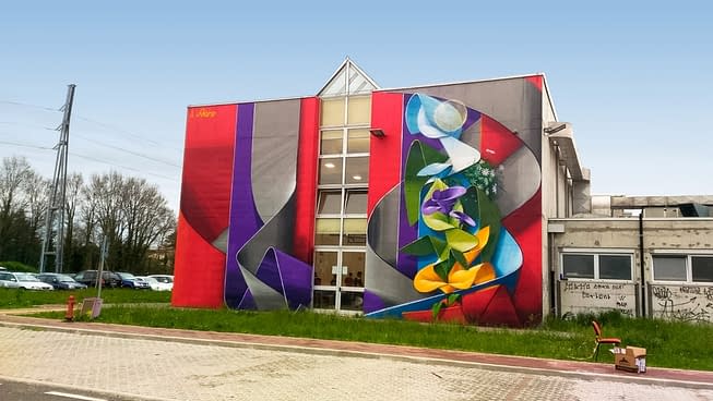 Lateral view of an high school building face with a mural painting depicting wide gray and purple ribbons composing the name Piero in a graffiti wildstyle decorated with yellow and purple flowers. Artwork by Dado.