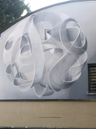 Main element of a mural painting depicting a spherical wildstyle graffiti forming the tag Dado colored in a very vanishing white and light gray tones.