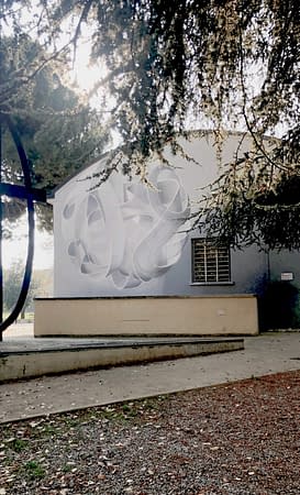 A wall behind trees with a mural painting depicting a spherical wildstyle graffiti forming the tag Dado colored in a very vanishing white and light gray tones.