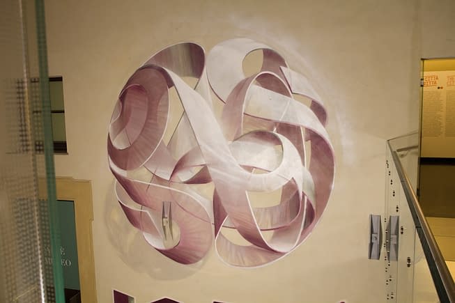 Mural painting inside a depicting a spherical wildstyle graffiti forming the tag Dado colored in a vanishing light pink and winy tones.