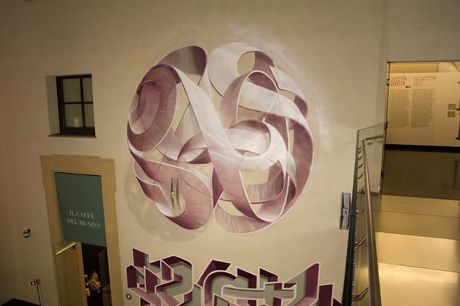 Total top view of a mural painting inside an art gallery depicting a spherical wildstyle graffiti forming the tag Dado colored in a vanishing light pink and winy tones.