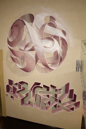 Protrait view of a mural painting on an indoor wall depicting a spherical wildstyle graffiti forming the tag Dado colored in a vanishing light pink and winy tones along with another lettering graffiti underneath it.