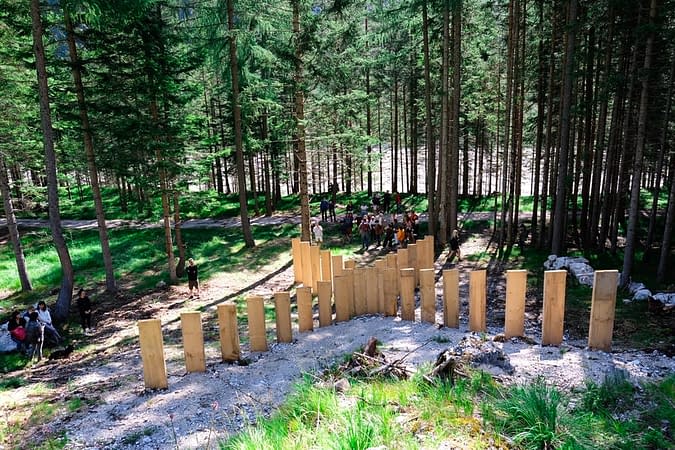 Back view of a wood sculpture composed from a series of tall wooden boards planted in the ground forming a big X just in the middle of a mountain path in a sunny spring day. A group of hikers are approaching the mountain path.
