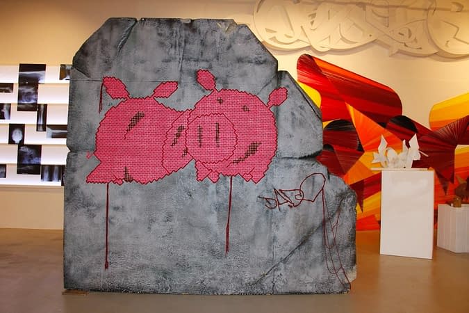 A big stone inside an art gallery, with two pig face characters stitch on it with wool in pink color with red outline, along with the tag Dado