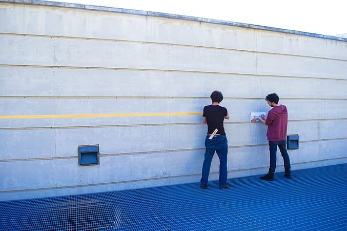 Two person preparing a long gray concrete wall to be used as a surface to expose drawings for an exhibition
