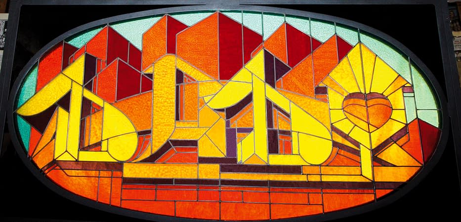 A stained glass window made colored glass pieces representing the word Dado in a graffiti style at the bottom of stylized skyscrapers.